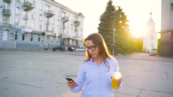 Mujer utiliza teléfono inteligente y beber jugo caminando por la calle — Vídeo de stock