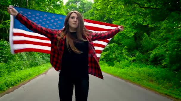 Jovem adolescente acenando uma bandeira dos EUA está patinando através da floresta. Movimento lento — Vídeo de Stock