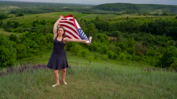 Una mujer ondeando una bandera de Estados Unidos está de pie en la cima de una colina - el concepto del Día de la Independencia de Estados Unidos — Vídeo de stock