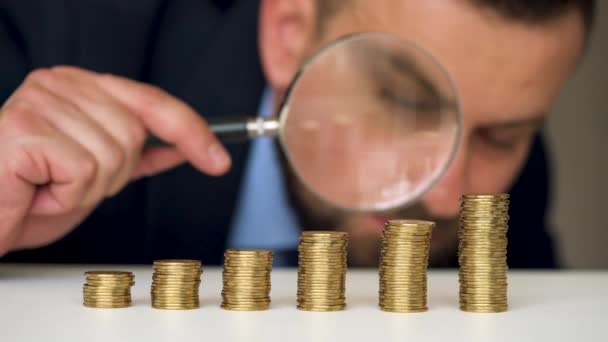 Businessman examines in a magnifying glass columns of coins — Stock Video