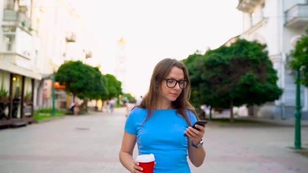 Женщина разговаривает по смартфону и пьет кофе, идя по улице. — стоковое видео