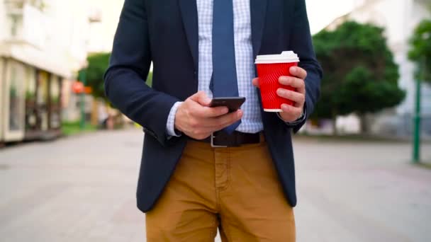 Чоловік використовує смартфон і п'є каву, що йде по вулиці — стокове відео