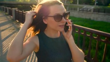 Kadın güneş gözlüğü smartphone üzerinde söz ederken kadar günbatımı, yakın sokakta yürüyen, yakın çekim. Ağır çekim