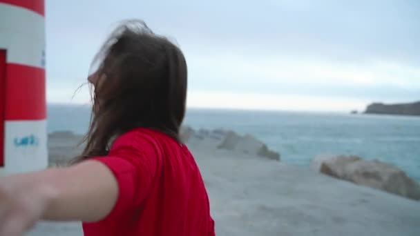 Следуйте за мной - счастливая молодая женщина в красном платье тянет парней рука об руку ходить к маяку на пляже на закате — стоковое видео