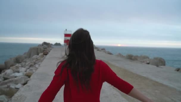 穿红衣服的女人在海洋海岸上运行到灯塔去 》。慢动作 — 图库视频影像