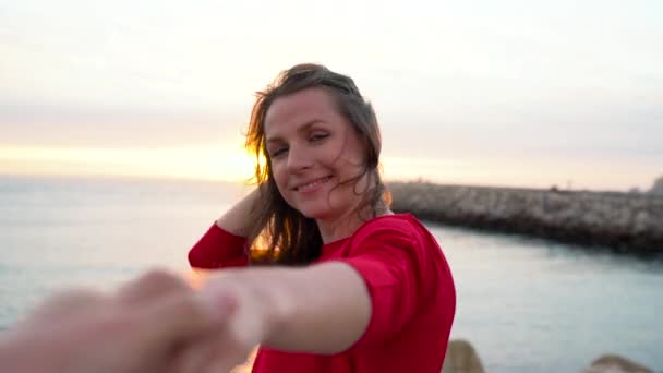 Suivez-moi - heureuse jeune femme en robe rouge tirant les gars main dans la main marchant vers le phare sur la plage au coucher du soleil — Video