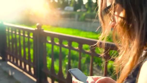 Женщина пользуется смартфоном во время прогулки по улице, на закате - вид сзади — стоковое видео