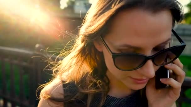 Mujer con gafas de sol hablando en el teléfono inteligente mientras camina por la calle al atardecer, de cerca, de cerca. Movimiento lento — Vídeos de Stock