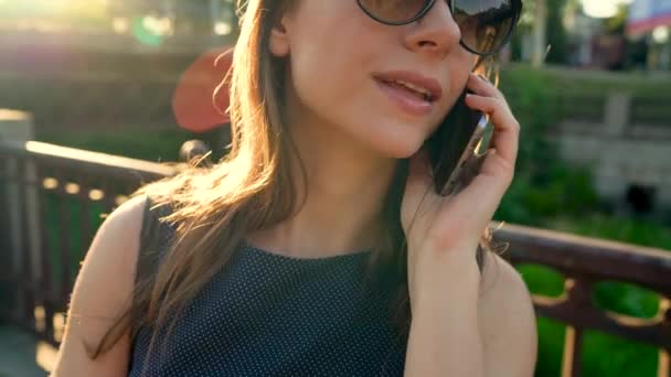 Kvinna i solglasögon talar på din smartphone medan gångavstånd ner på gatan i solnedgången, nära upp, närbild — Stockvideo