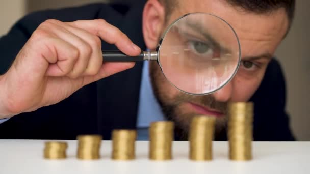 Businessman examines in a magnifying glass columns of coins — Stock Video