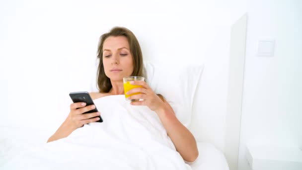 Mujer linda comprueba el teléfono inteligente y bebe jugo de naranja en la cama por la mañana — Vídeos de Stock