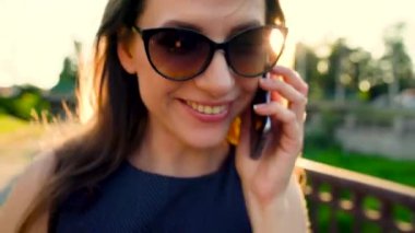 Kadın güneş gözlüğü smartphone üzerinde söz ederken kadar günbatımı, yakın sokakta yürüyen, yakın çekim