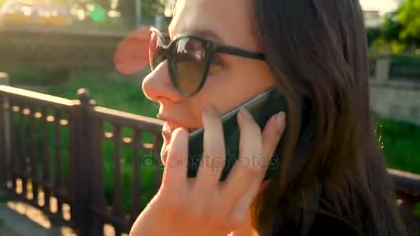 Жінка в сонцезахисних окулярах говорить по смартфону під час прогулянки по вулиці на заході сонця - вид ззаду, крупним планом — стокове відео