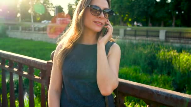 Mujer con gafas de sol hablando en el teléfono inteligente mientras camina por la calle al atardecer, de cerca, de cerca — Vídeo de stock