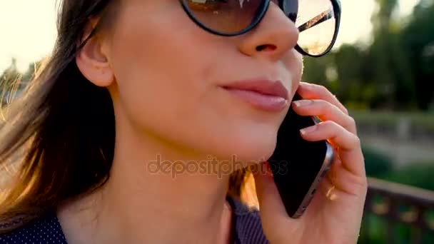 Жінка в сонцезахисних окулярах говорить по смартфону під час прогулянки по вулиці на заході сонця, крупним планом — стокове відео