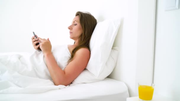 Femme mignonne vérifie le smartphone et boit du jus d'orange au lit le matin — Video