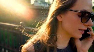 Kadın güneş gözlüğü smartphone üzerinde söz ederken kadar günbatımı, yakın sokakta yürüyen, yakın çekim