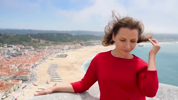 Mulher de vestido vermelho relaxante no topo de uma colina acima da cidade de Nazaré, Portugal, câmera lenta — Vídeo de Stock