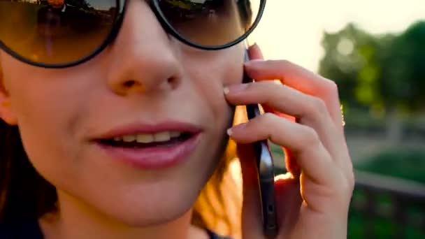 Женщина в солнечных очках разговаривает по смартфону во время прогулки по улице на закате, крупным планом — стоковое видео