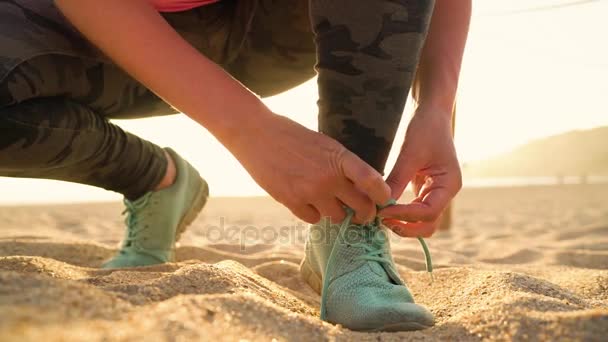 Взуття для бігу - жінка прив'язує взуття мереживами на піщаному пляжі під час заходу сонця. Повільний рух — стокове відео
