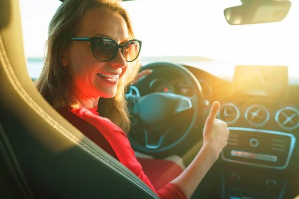 Ευτυχισμένη γυναίκα με κόκκινο φόρεμα με τον αντίχειρα επάνω να κάθεται στο αυτοκίνητο — Φωτογραφία Αρχείου