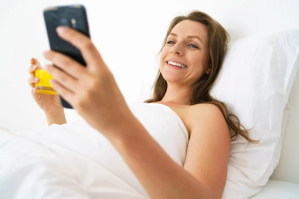 Carina donna controlla lo smartphone e beve succo d'arancia a letto — Foto Stock