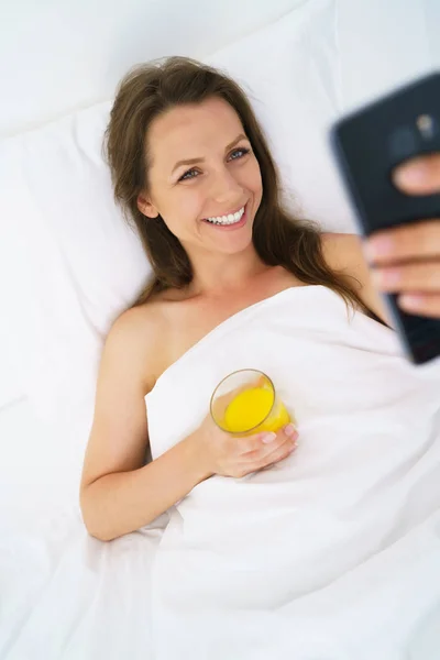 Carina donna controlla lo smartphone e beve succo d'arancia a letto — Foto Stock