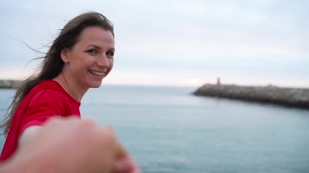 Folgen Sie mir - glückliche junge Frau in rotem Kleid zieht Jungs Hand in Hand zum Leuchtturm am Strand bei Sonnenuntergang. — Stockvideo