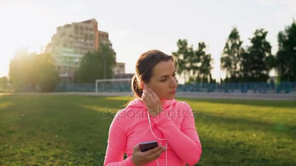 3 in 1 video. Vrouw met hoofdtelefoon en smartphone loopt door het stadion bij zonsondergang — Stockvideo