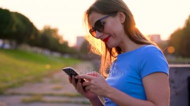 Kadın bir akıllı telefon açık havada günbatımında kullanarak güneş gözlüğü