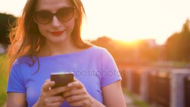 Kadın bir akıllı telefon açık havada günbatımında kullanarak güneş gözlüğü