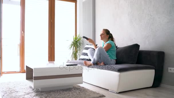 Jeune, belle femme allongée sur le canapé et regardant la télé. Elle allume la télécommande indéfiniment, traquant le tir — Video