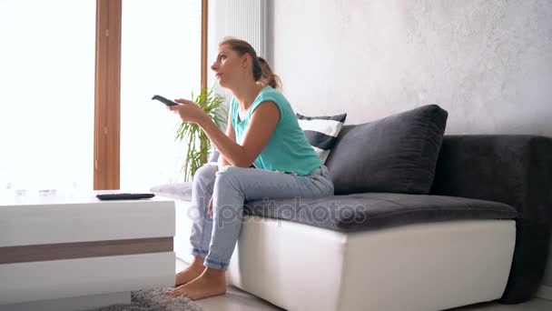 Молодая, красивая женщина лежит на диване и смотрит телевизор. Она включает пульт бесконечно, отслеживая выстрел — стоковое видео