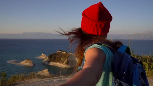 Siga-me - jovem mulher feliz em um chapéu vermelho e com uma mochila nas costas puxando a mão caras. Caminhando de mãos dadas até o Cabo Drastis na ilha de Corfu, na Grécia — Vídeo de Stock