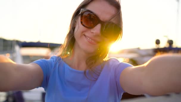 Женщина в солнечных очках делает селфи на фоне пристани с большим количеством яхт и лодок на закате, крупным планом. Медленное движение — стоковое видео