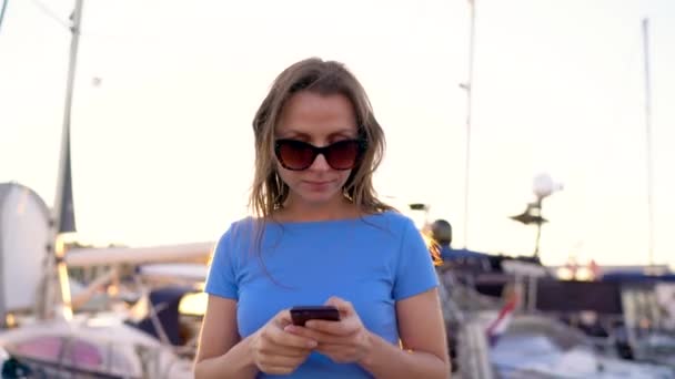 Femme en lunettes de soleil en utilisant un smartphone tout en marchant sur un quai avec beaucoup de yachts et bateaux au coucher du soleil, gros plan, gros plan 