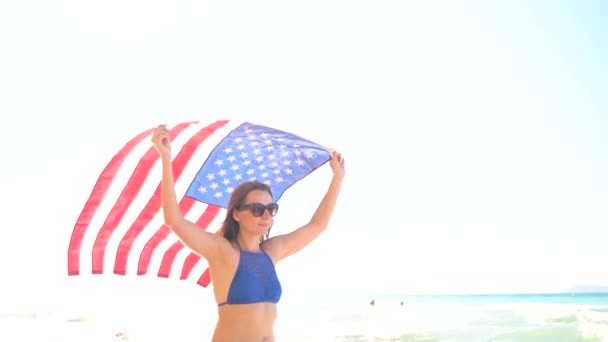 Bikini de plage femme avec drapeau américain marche insouciante le long de l'eau sur la plage. Concept de fête de l'indépendance États-Unis — Video
