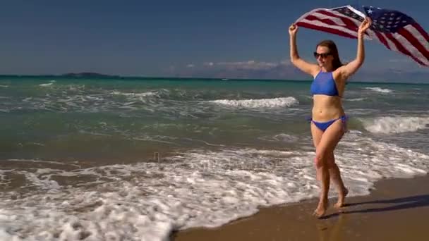 Strand bikini vrouw met ons vlag loopt langs het water op het strand. Concept van Independence Day Usa. Slow motion — Stockvideo