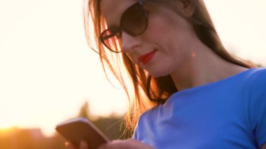Kadın bir akıllı telefon açık havada günbatımında kullanarak güneş gözlüğü