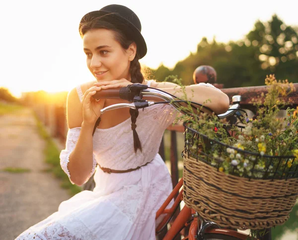सूर्याच्या वेळी फुलांसह तिच्या सायकलवर बसलेली तरुण सुंदर महिला — स्टॉक फोटो, इमेज