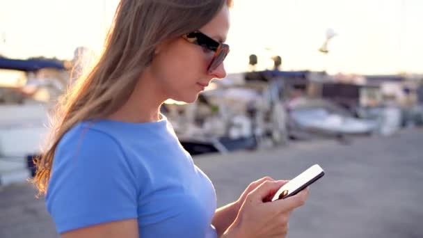 Femme en lunettes de soleil parlant sur le smartphone tout en marchant sur un quai avec beaucoup de yachts et bateaux au coucher du soleil, gros plan — Video