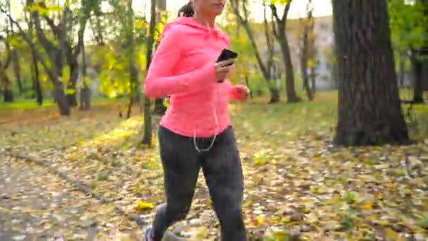 Женщина с наушниками и смартфоном бегает по осеннему парку на закате — стоковое видео