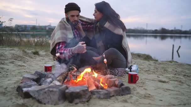 Pareja enamorada sentada alrededor de la hoguera en el lago, hablando y bebiendo té caliente — Vídeos de Stock
