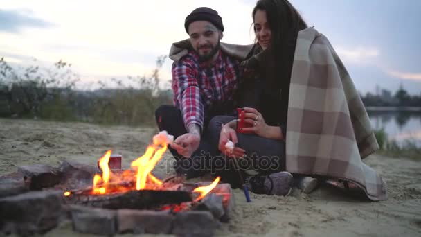 Pareja enamorada sentada alrededor de la hoguera en el lago, hablando, bebiendo té caliente y malvaviscos fritos — Vídeos de Stock