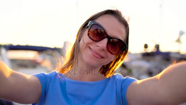 Mulher de óculos de sol faz selfie no fundo da marina com um monte de iates e barcos ao pôr do sol, close up. Movimento lento — Vídeo de Stock