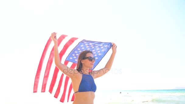 Praia mulher biquíni com bandeira dos EUA despreocupado andando ao longo da água na praia. Conceito de Dia da Independência EUA — Vídeo de Stock