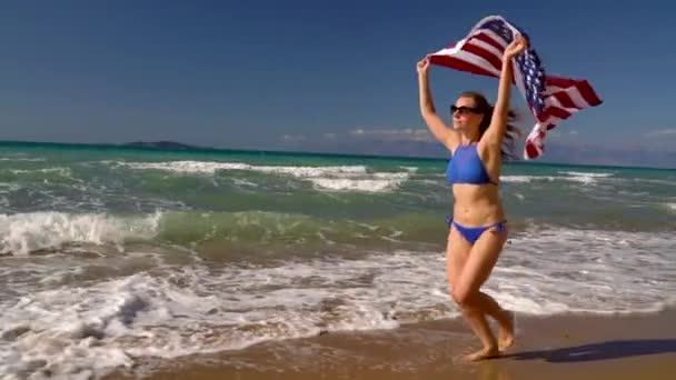 Strand bikini nő velünk zászló fut a víz mentén a tengerparton. Fogalma az Usa a függetlenség napja. Lassú mozgás — Stock videók