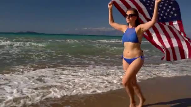 Mujer bikini de playa con bandera de Estados Unidos corriendo a lo largo del agua en la playa. Concepto del Día de la Independencia USA — Vídeo de stock