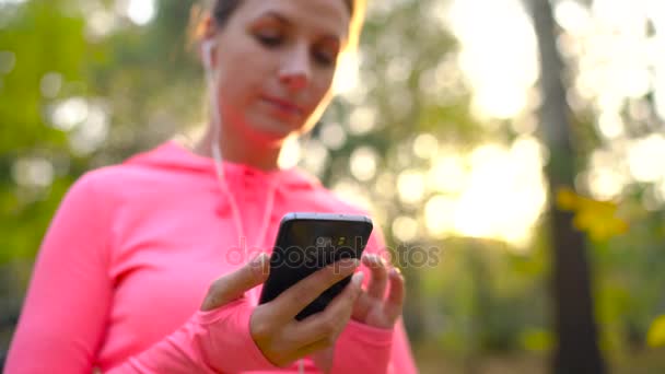 Mujer con auriculares y un smartphone elige la música para correr por el parque de otoño — Vídeo de stock