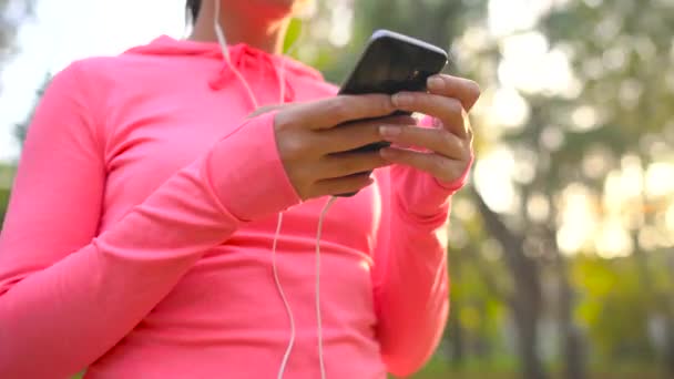 Женщина в наушниках и смартфоне выбирает музыку для пробежки по осеннему парку — стоковое видео